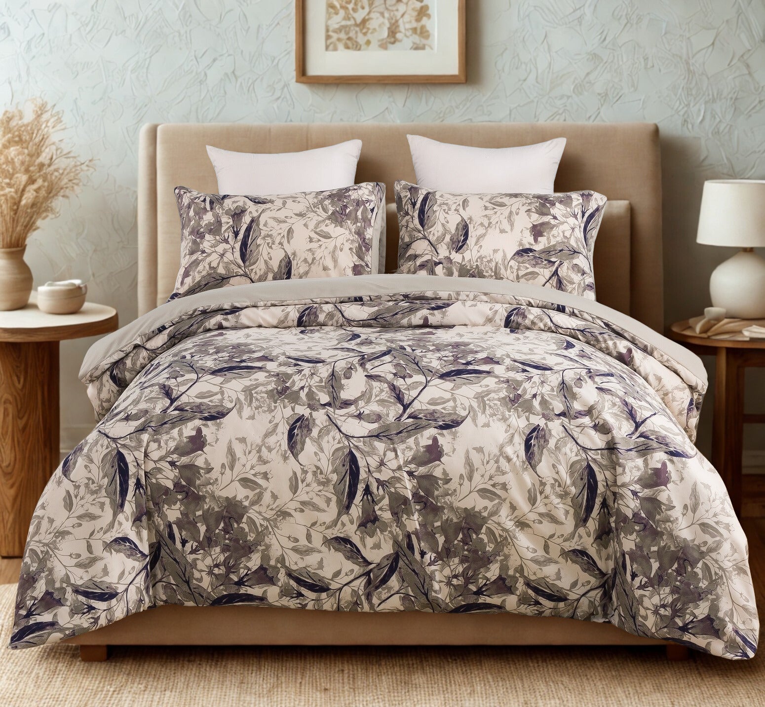 A serene bedroom features a bed with a duvet cover and pillows showcasing a delicate gray and white floral pattern. The bed is flanked by two wooden nightstands, one with a lamp and a small bowl, and the other with a vase of dried branches. A framed botanical print hangs on the light blue textured wall, and a woven rug covers the floor, adding warmth to the space