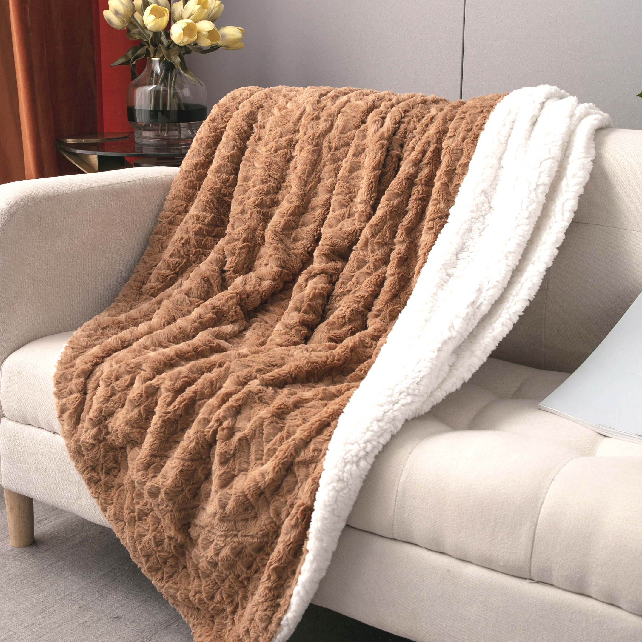 A cozy beige loveseat with a plush cognac brown faux fur blanket draped over it, featuring a soft white sherpa lining. The scene is set against a light gray wall with sunlight streaming in, creating a warm and inviting atmosphere, ideal for home decor and relaxation