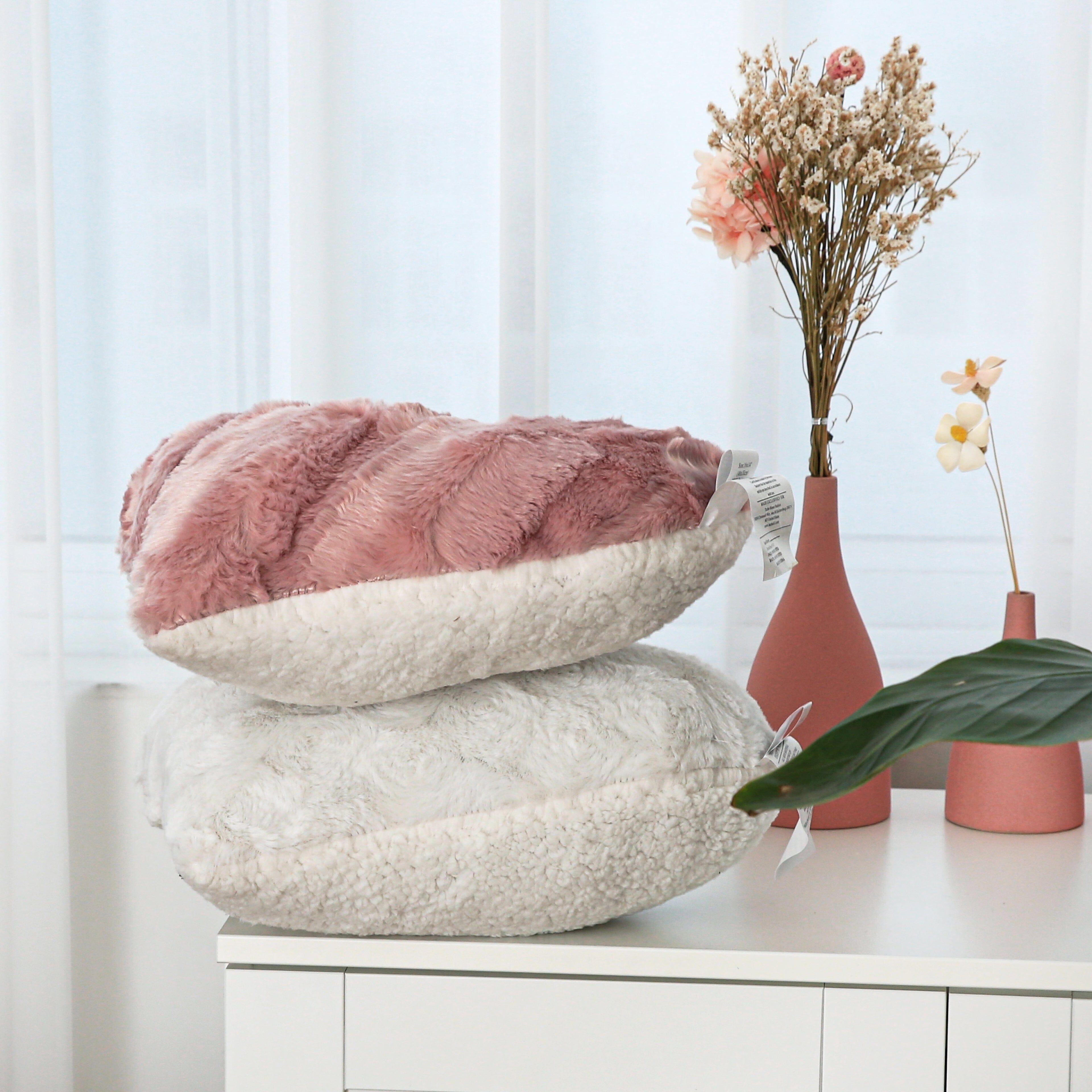 A stack of decorative pillows in various textures and patterns sits on a wooden dresser. A vase filled with pink and purple flowers is positioned to the right