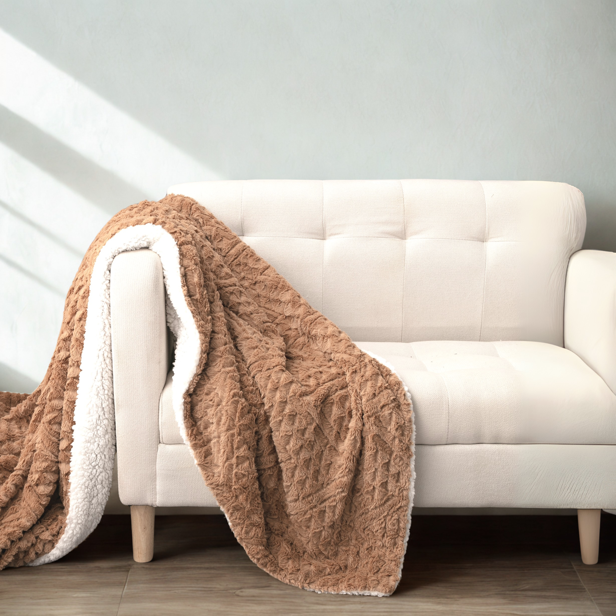 A cozy beige loveseat with a plush brown faux fur blanket draped over it, featuring a soft white sherpa lining. The scene is set against a light gray wall with sunlight streaming in, creating a warm and inviting atmosphere, ideal for home decor and relaxation