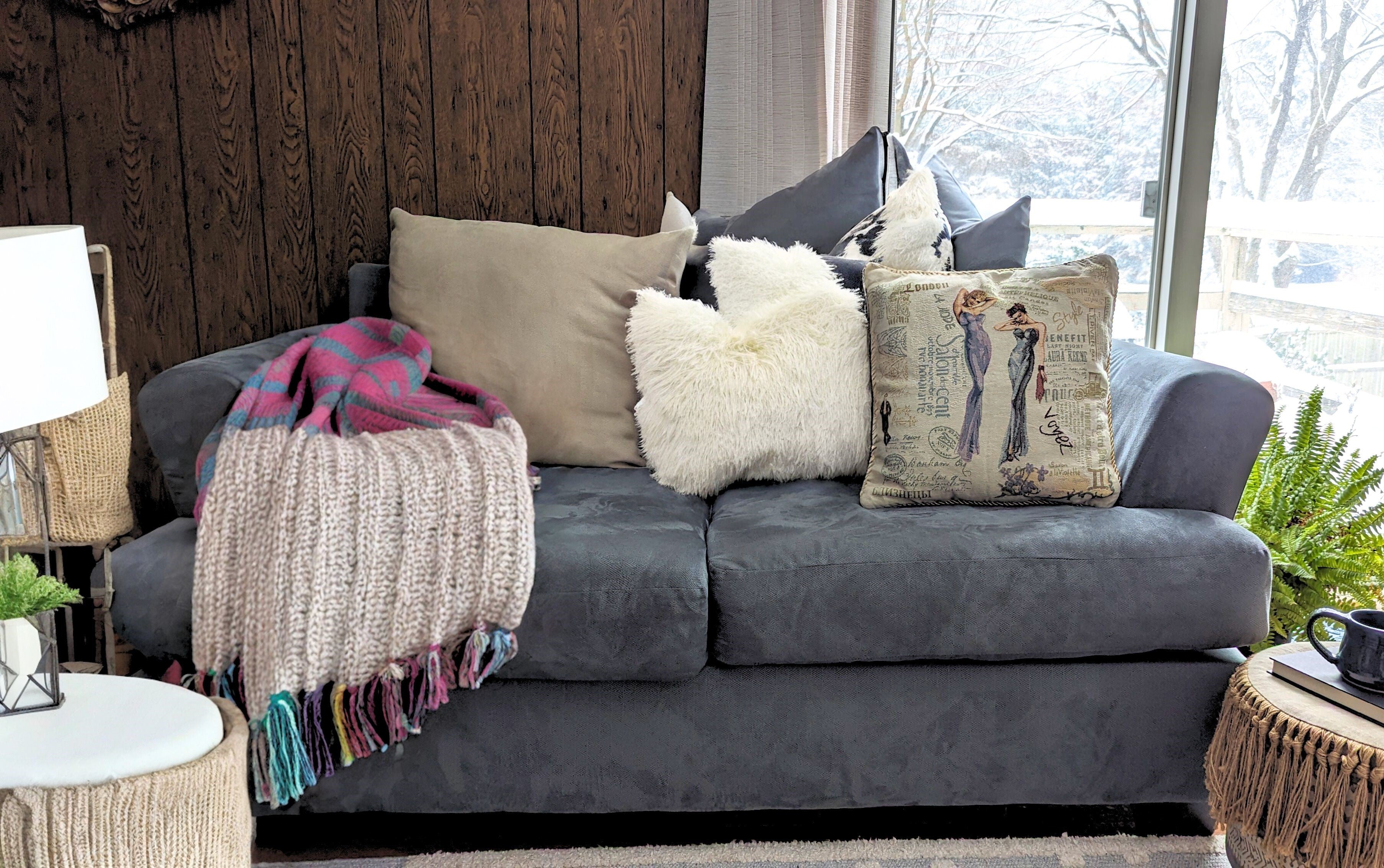 A living room scene with a comfortable couch adorned with decorative pillows and a knitted throw blanket. The tapestry accent pillow has a pattern of 2 girls on it