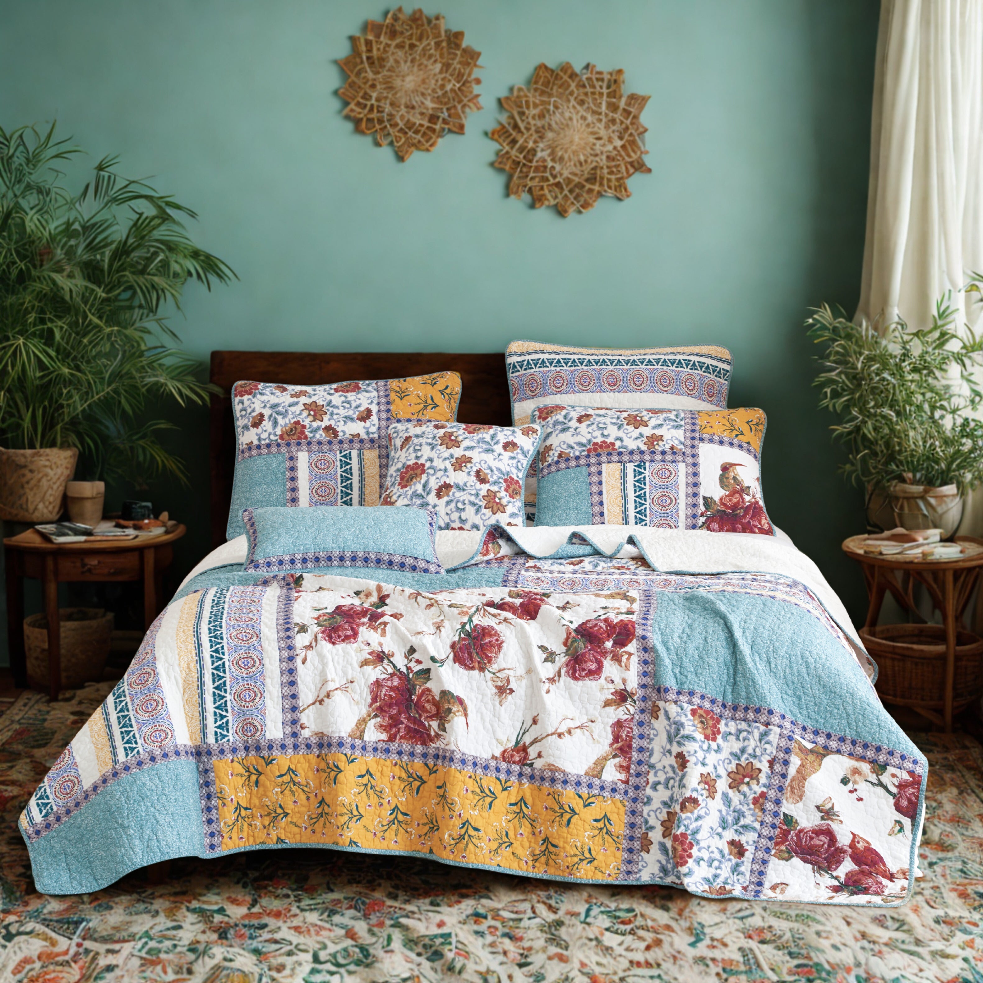 A vibrant bedroom features a bed with a patchwork quilted bedspread quilt bedding in blue, yellow, and white, adorned with red floral and geometric patterns. The bed is flanked by two wooden nightstands, one with a lamp and small items, and the other with a plant and books. The room has teal walls decorated with two woven wall hangings, a large window with sheer curtains, and a colorful patterned rug on the floor, with potted plants adding a touch of greenery