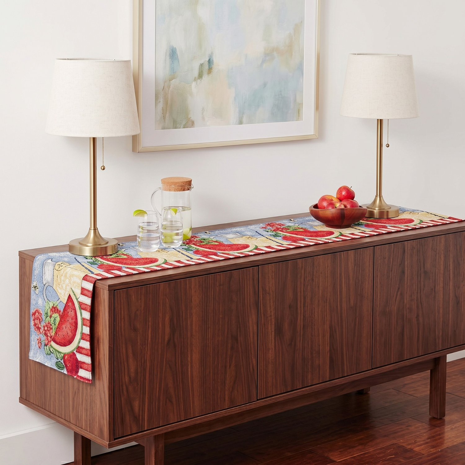 Tache woven tapestry table runner with a summer watermelon and lemonade design, displayed on a wooden sideboard with a bowl of fresh apples and two table lamps.