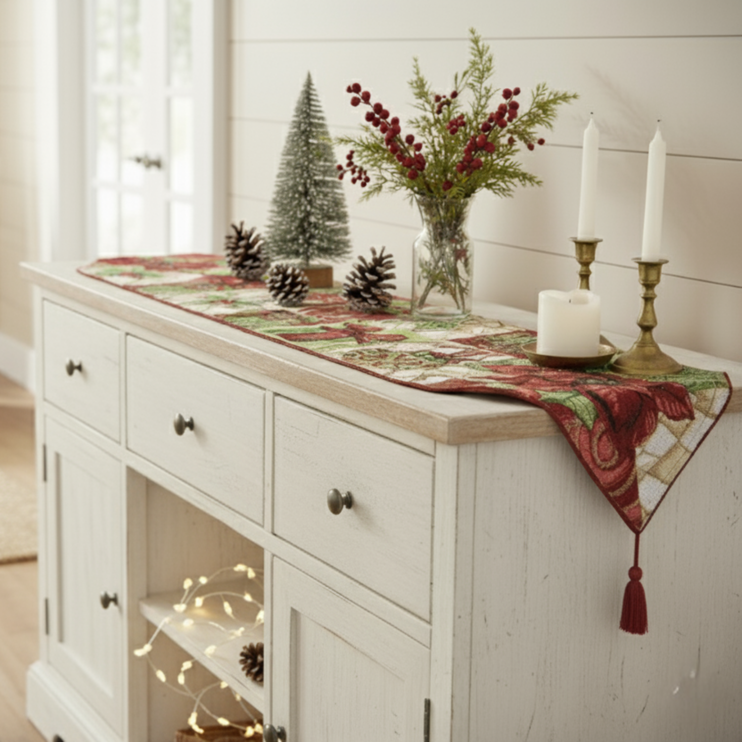 Decorative table setting with a red and green runner, candles, and pinecones on a white cabinet.