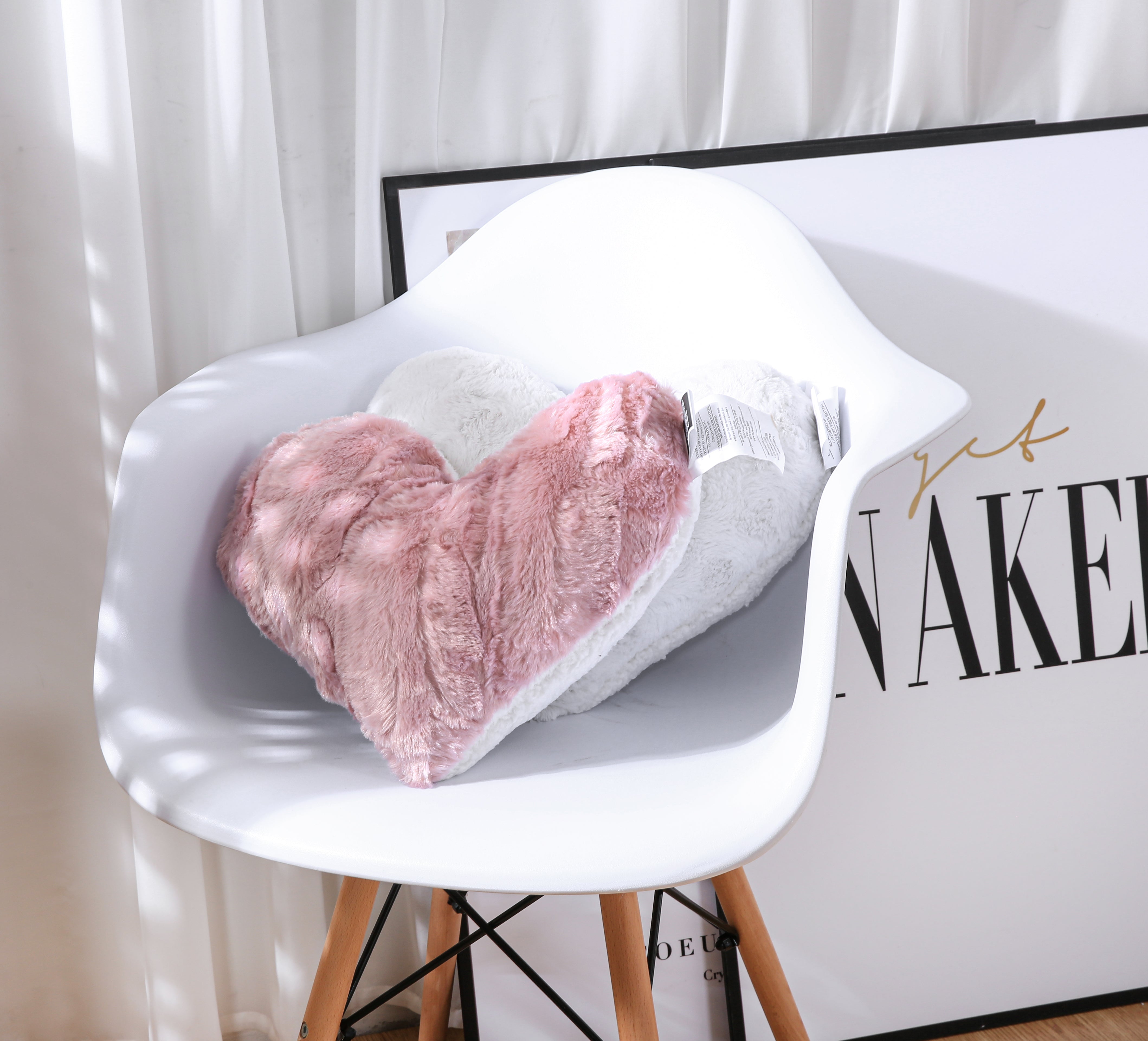A close-up view of two plush faux fur heart-shaped pillows nestled on a white armchair. The pillows are soft and textured, resembling faux fur. One is a pale blush dusty rose pink color, while the other is white. They rest on a white armchair with a simple, modern design.