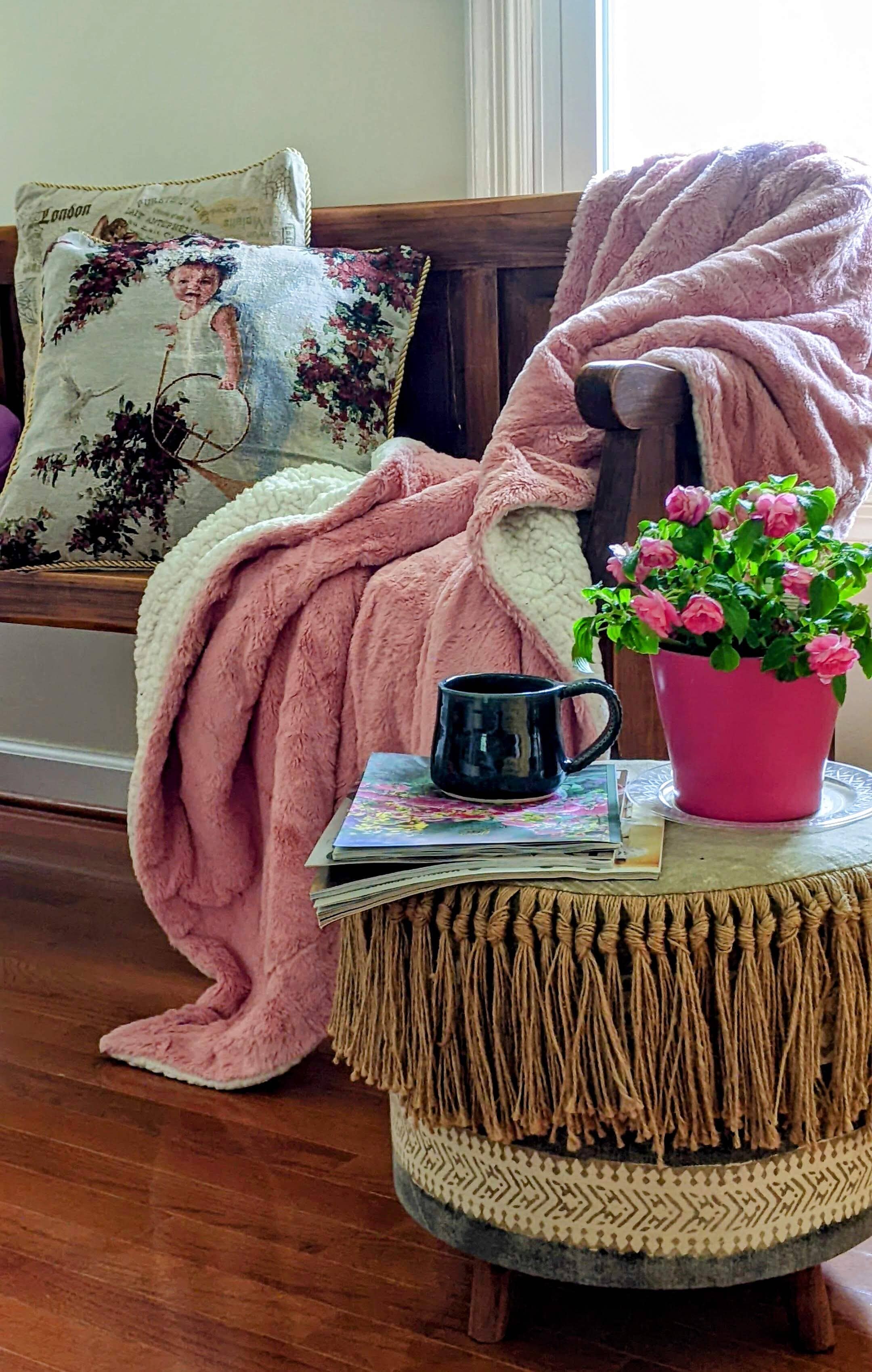 A soft pink blanket draped over a chair next to an ottoman with magazines and a cup of coffee on it