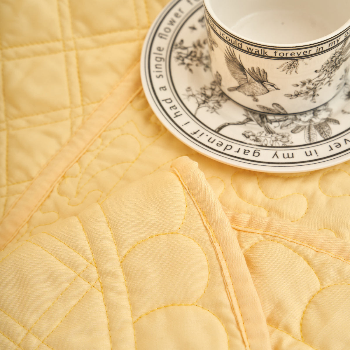 Yellow quilted throw blanket with scalloped border styled flat beside a botanical teacup and saucer